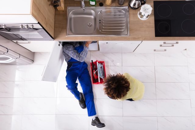 Plumber fixing under-sink pipes while homeowner watches